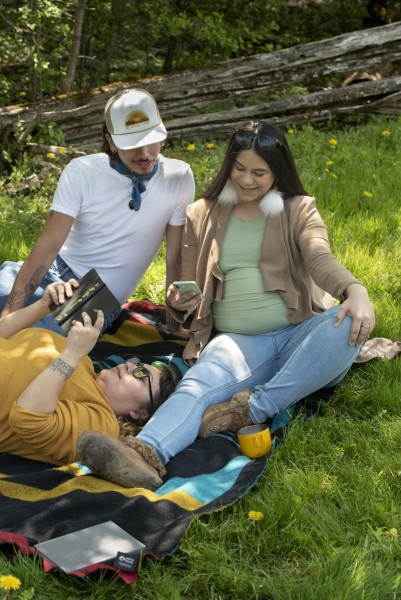 Friends having a picnic and looking at social media while their other friend reads